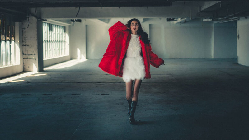 Model walking towards camera wearing a red coat and white dress