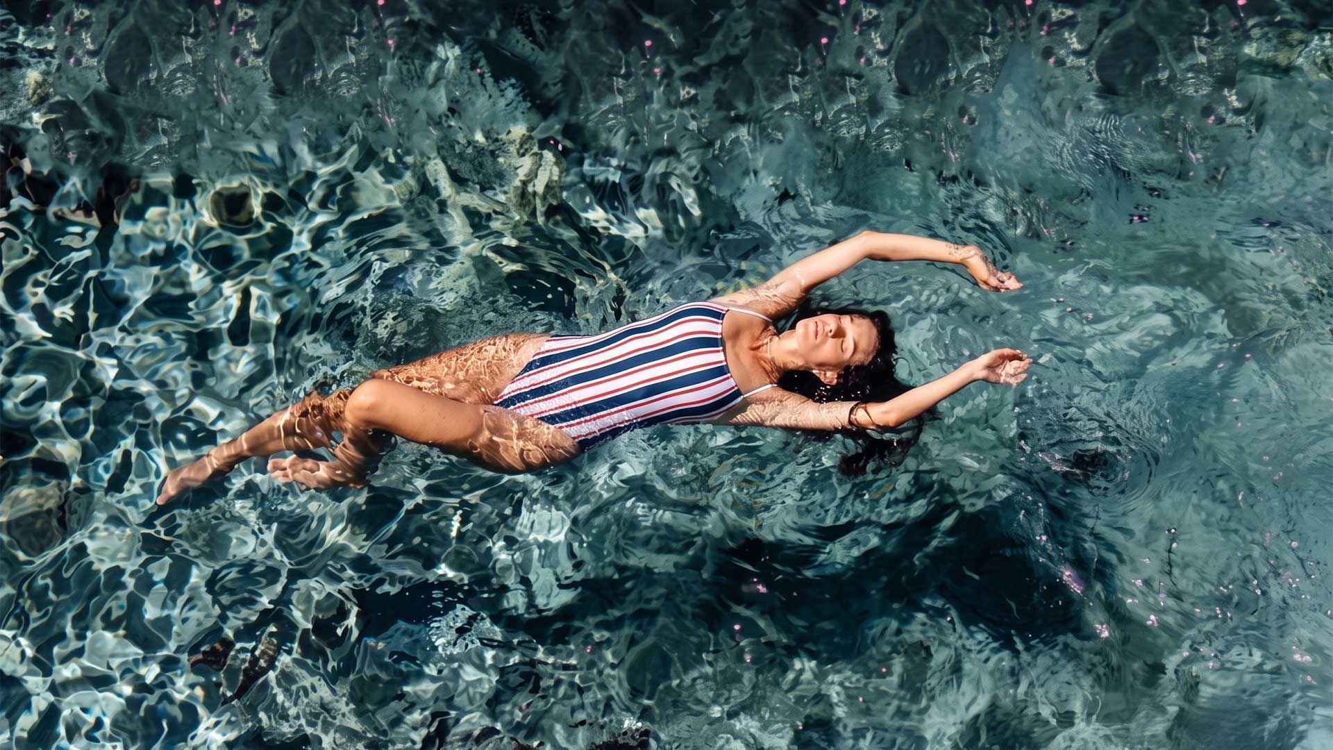 woman floats on her back in clear turquoise water, wearing a red, white, and blue striped swimsuit.