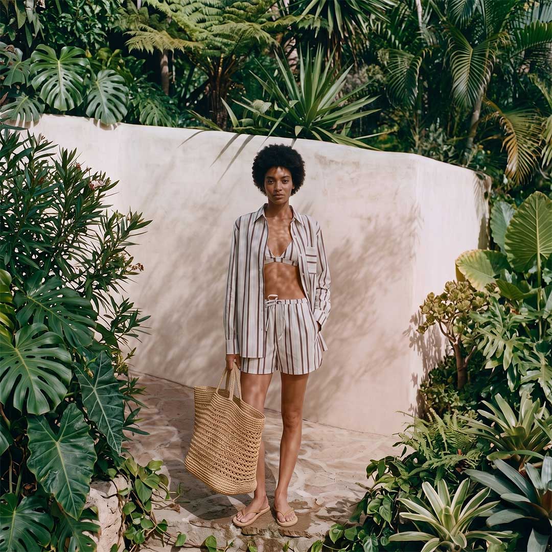 woman in a striped beige outfit (button-down shirt open over a bralette and striped shorts) standing in a lush tropical garden, holding a woven straw tote and wearing flip-flop sandals.