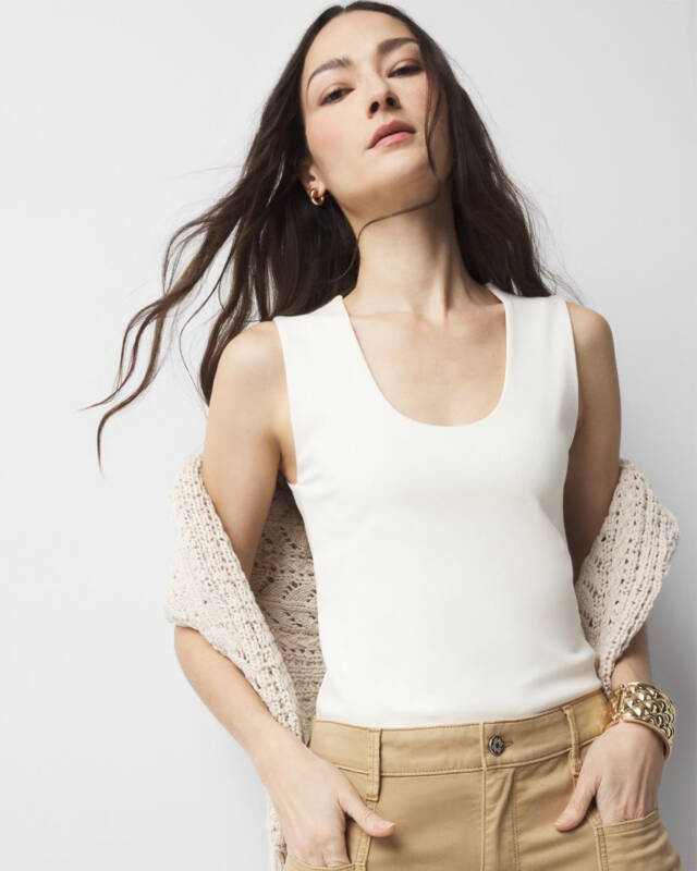 model wearing a white sleeveless top tucked into beige high-waisted pants, with a light knit cardigan draped over shoulders and gold jewelry