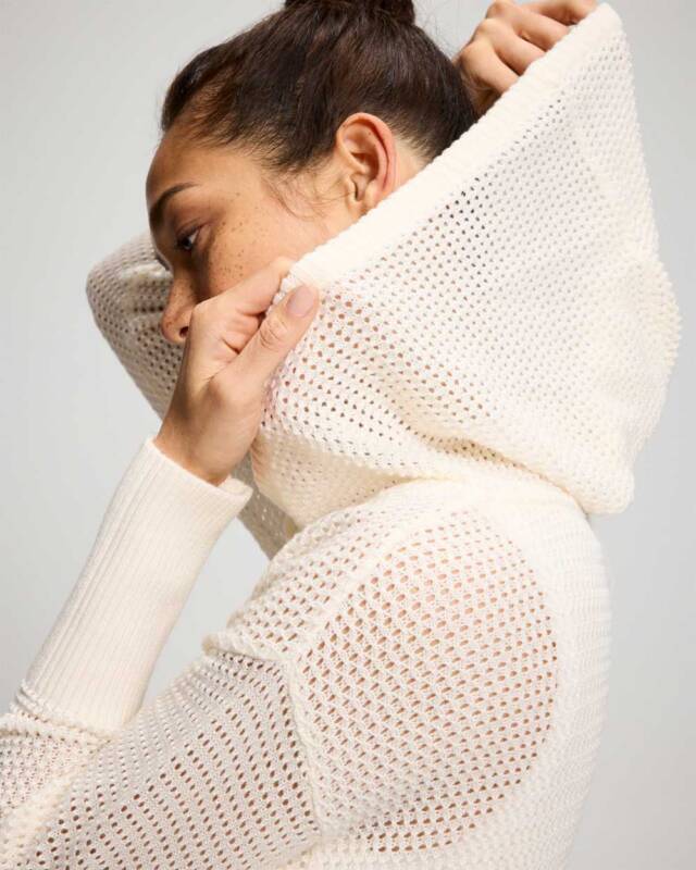 side view of a person with dark hair in a cream knit hoodie, adjusting the hood.