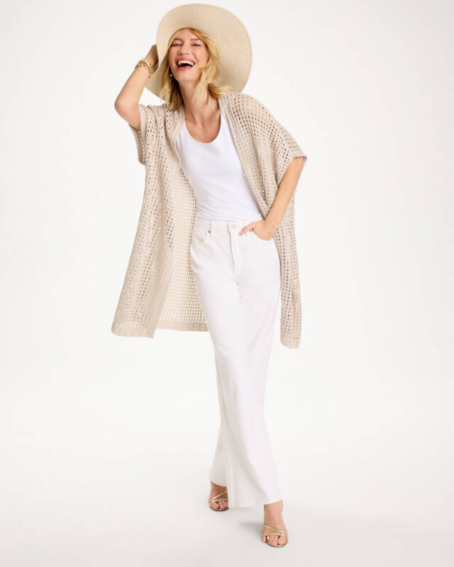 woman wearing a wide-brim straw hat, white tank top, beige crocheted cardigan, and white wide-leg pants, posing and smiling playfully.