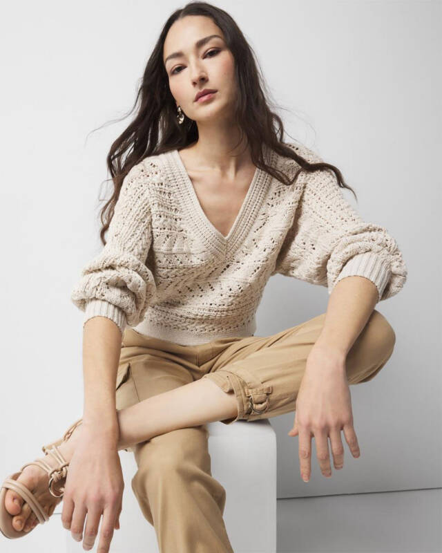 woman seated on a white block wearing a cream knit sweater and beige cargo pants, posing for a fashion shoot.