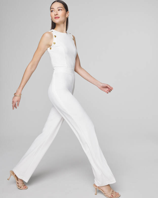 woman in a sleeveless white jumpsuit with gold button details walking against a gray studio backdrop.