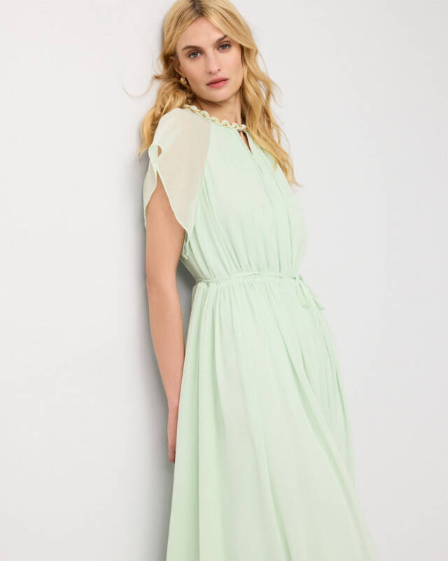 model wearing a pale mint-green maxi dress with sheer sleeves, standing against a light wall and looking at the camera.