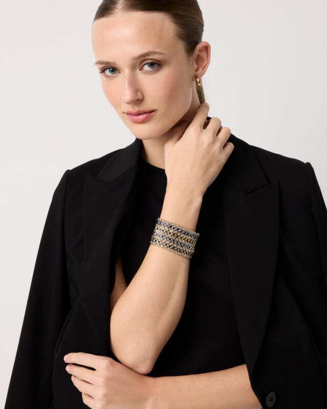 model wearing a black blazer with a wide blue-and-gold bracelet, posed with her hand near her neck and looking at the camera.