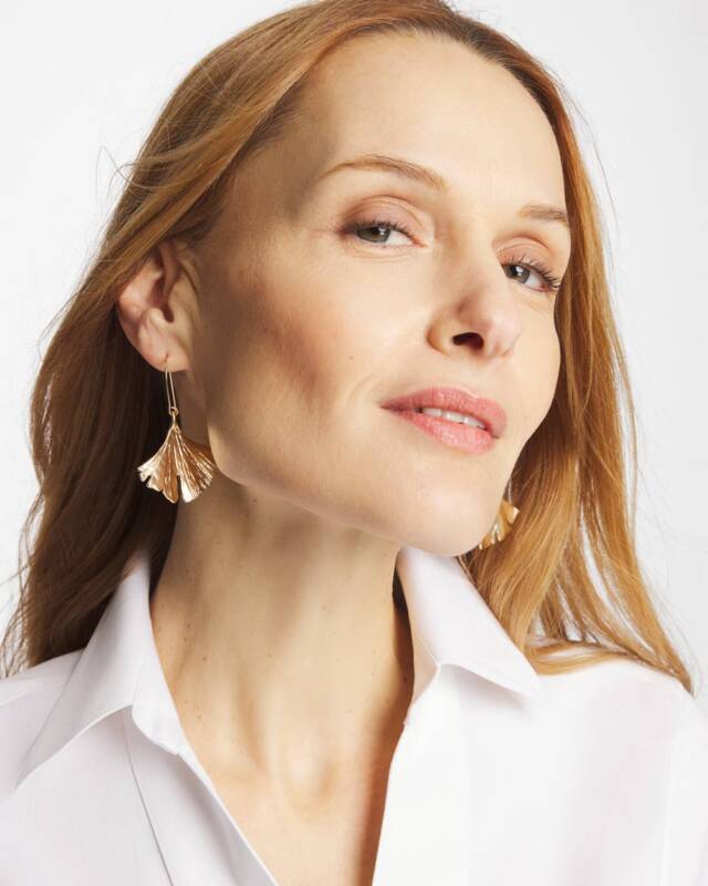 close-up of a woman with reddish hair wearing gold fan-shaped earrings and a white collared shirt, showing the jewelry detail.