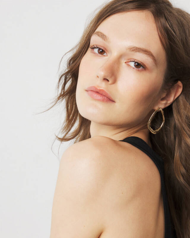 close-up portrait of a woman with brown hair, wearing a black strap top and gold teardrop hoop earrings, looking over her shoulder.
