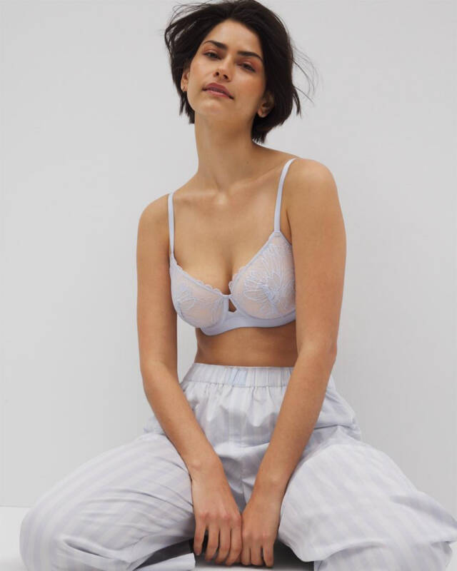 woman modeling a pale blue lace bra with light striped pajama pants, seated against a neutral background.