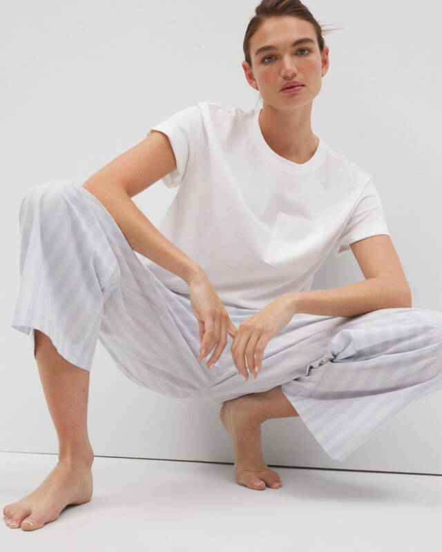 woman wearing a white t-shirt and light striped pajama pants crouches barefoot against a white backdrop, looking at the camera.