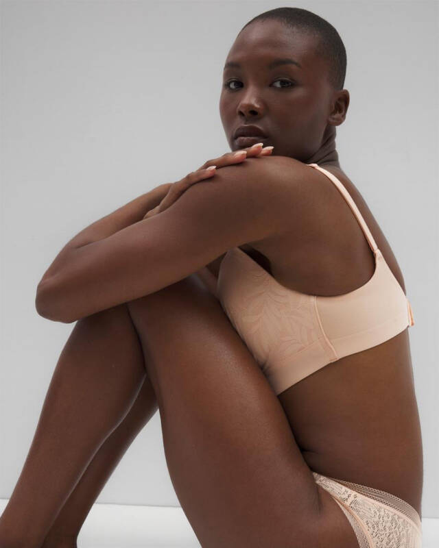 woman with a shaved head sits with knees drawn up, wearing nude lingerie and gazing at the camera against a gray background.