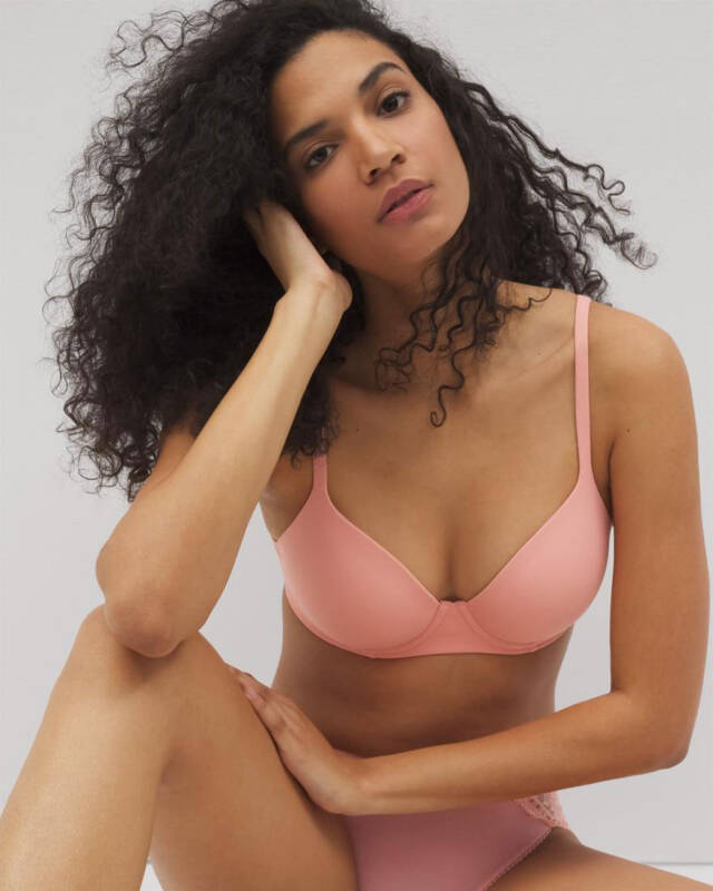 woman with curly hair wearing a pink bra and matching panties, seated against a light gray background.