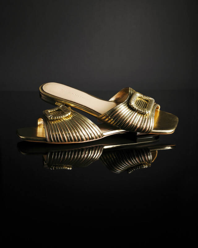 gold metallic slide sandals with pleated, ribbed straps resting on a black mirror-like surface.
