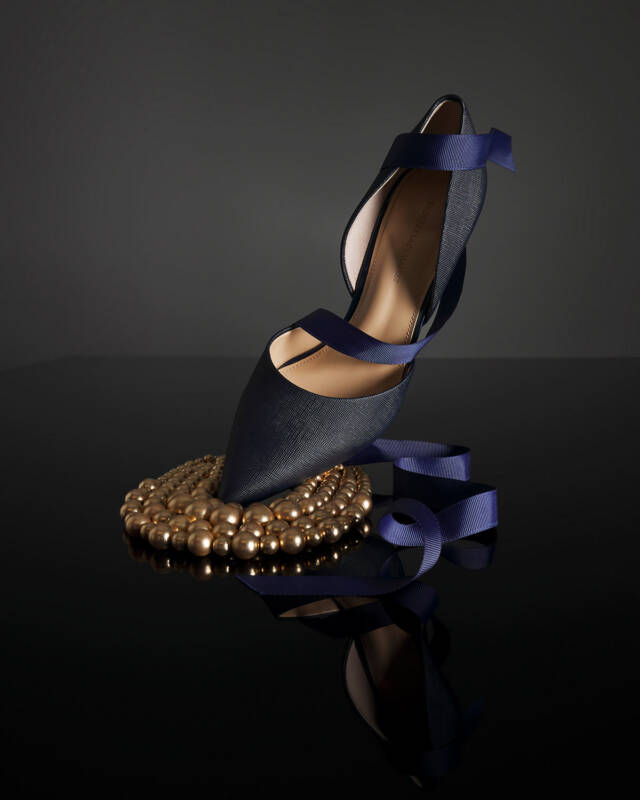 navy blue pointed-toe high heel with ribbon straps resting on a gold bead necklace on a glossy black surface.