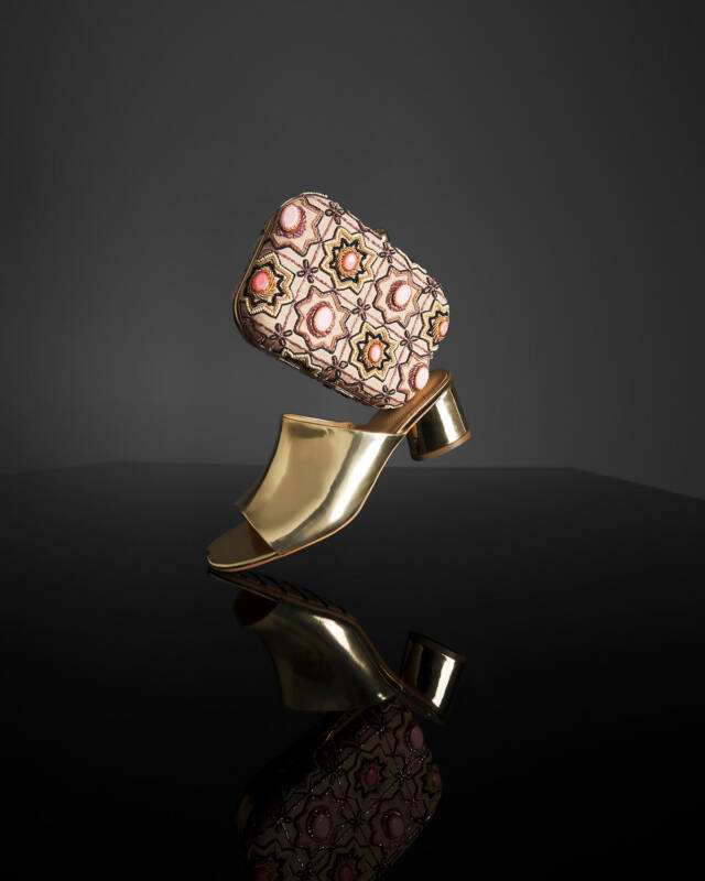 gold metallic high heel with an embellished patterned clutch resting on its toe against a dark studio backdrop.