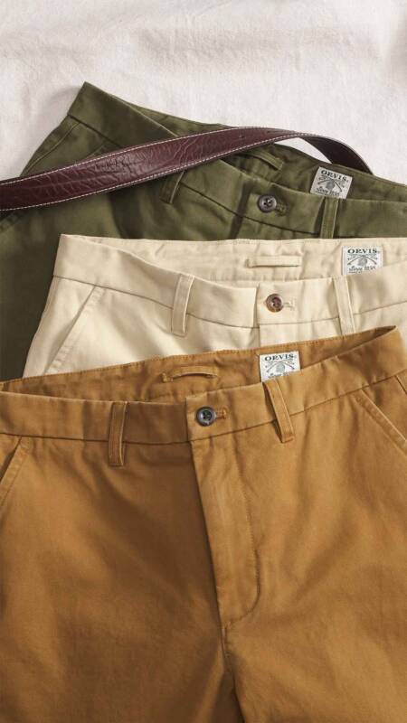 stacked chinos in olive, beige, cream, and camel with a brown leather belt draped across the top, showing waistband details and tags (orvis).