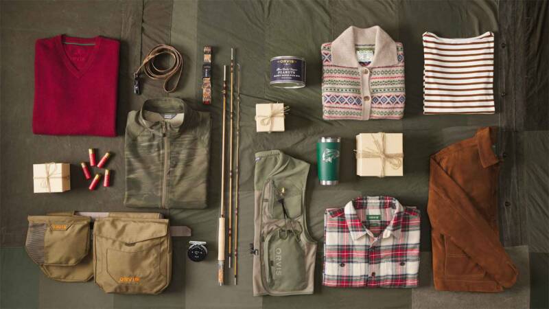 top-down layout of outdoor gear and clothing arranged on a dark tarp, including jackets, shirts, backpacks, and accessories.