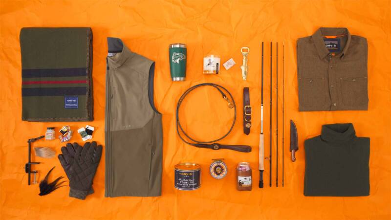 flat lay of outdoor gear on an orange background: jacket, fleece, blanket, belt, fishing rods, canteen, gloves, and small accessories arranged neatly.